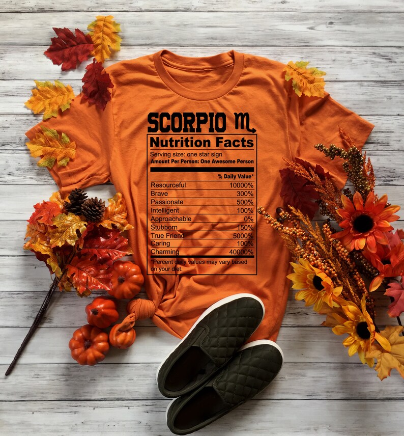 May include: Orange t-shirt with a black graphic that reads "SCORPIO M. Nutrition Facts" with a list of personality traits and percentages. The t-shirt is surrounded by fall leaves and pumpkins.