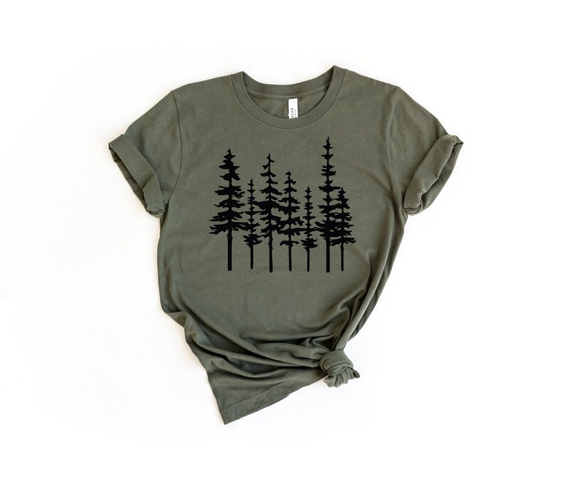 May include: Olive green t-shirt featuring a black graphic of stylized evergreen trees. The short-sleeved shirt is made of soft material and has rolled-up sleeves. The shirt is displayed on a white background.