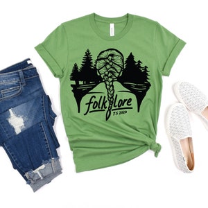 May include: A green t-shirt with the word "folklore" and a silhouette of a braided hairstyle and trees. The shirt is paired with blue jeans and white slip-on shoes.