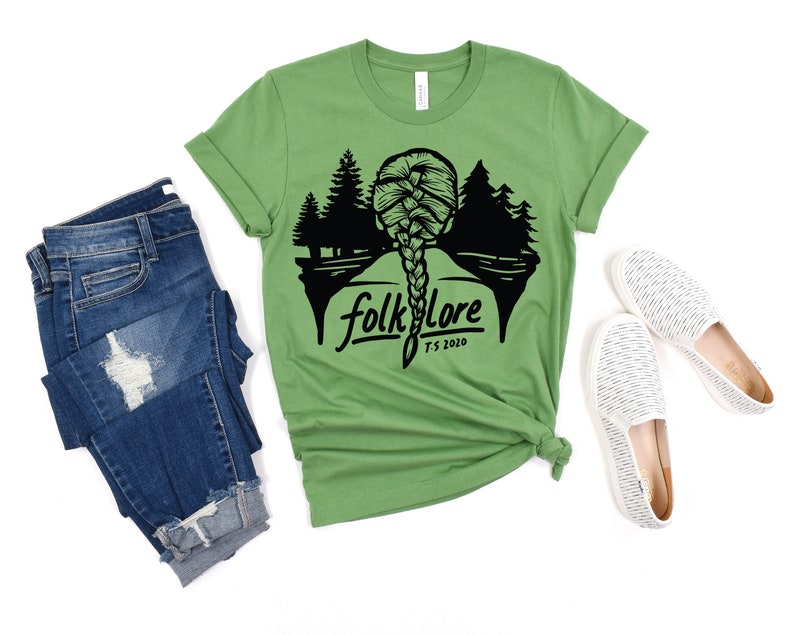 May include: A green t-shirt with the word "folklore" and a silhouette of a braided hairstyle and trees. The shirt is paired with blue jeans and white slip-on shoes.