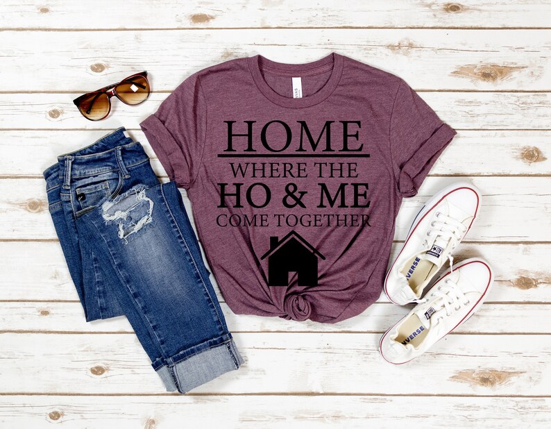 May include: A maroon t-shirt with the text "HOME WHERE THE HO & ME COME TOGETHER" in black lettering. A small house icon is below the text. The shirt is laid flat on a white wooden surface with a pair of jeans and white sneakers.
