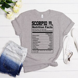 May include: Gray t-shirt with a black graphic of a nutrition facts label for Scorpio. The label lists traits like resourceful, brave, passionate, intelligent, approachable, stubborn, true friend, caring, and charming with percentages next to each trait.
