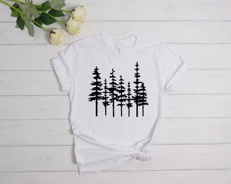 May include: White t-shirt featuring a black graphic of several stylized pine trees. The trees are depicted with detailed silhouettes, creating a nature-inspired design. The shirt is made of a soft material and has a classic crew neck.