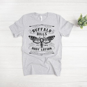 May include: Gray t-shirt with a black and white graphic of a moth with a skull on its body. The text reads "It rubs the lotion on its Buffalo Bills. Comes with its own body lotion. Skin or else it gets the hose again."