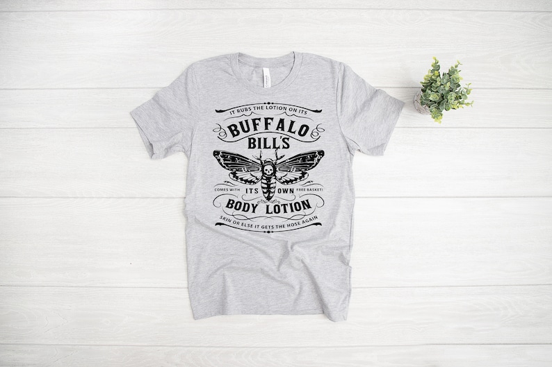 May include: Gray t-shirt with a black and white graphic of a moth with a skull on its body. The text reads "It rubs the lotion on its Buffalo Bills. Comes with its own body lotion. Skin or else it gets the hose again."