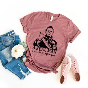 K&ouml;nnte beinhalten: Ein mauvefarbenes T-Shirt mit einer schwarzen Grafik eines Mannes, der ein Messer h&auml;lt, und dem Text "A real man will chase after you". Das Shirt wird mit einer blauen Jeans und rosa Sneakern kombiniert.