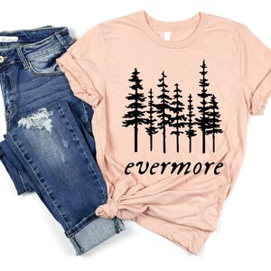 May include: A peach-colored t-shirt with a black graphic of stylized pine trees and the word "evermore" in a script font. The shirt is paired with blue denim jeans.