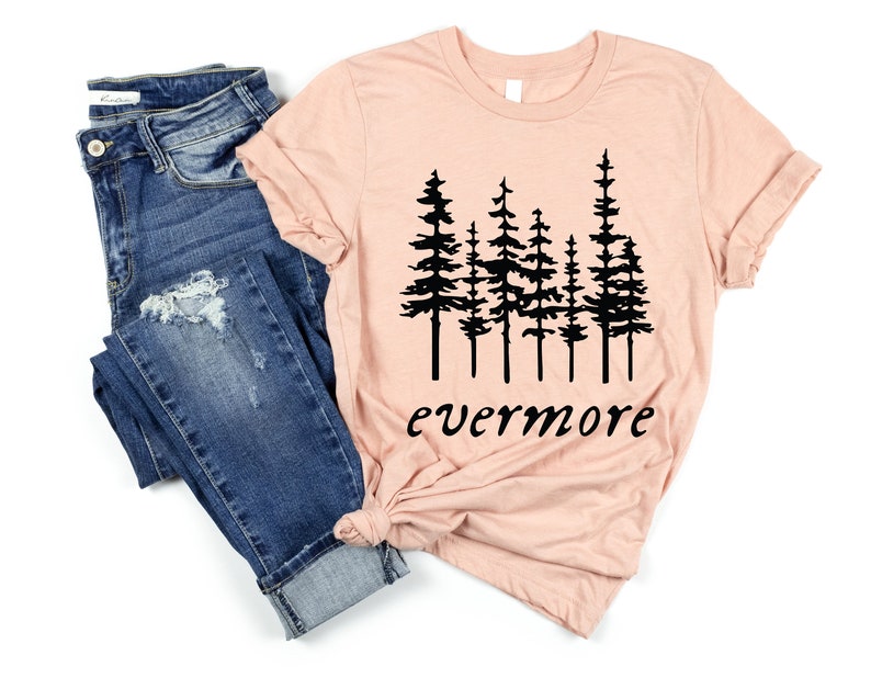 May include: A peach-colored t-shirt with a black graphic of stylized pine trees and the word "evermore" in a script font. The shirt is paired with blue denim jeans.