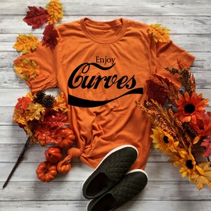 May include: Orange t-shirt with the text "Enjoy Curves" in a black retro font. The shirt is laid out on a white wooden surface with fall foliage and pumpkins.