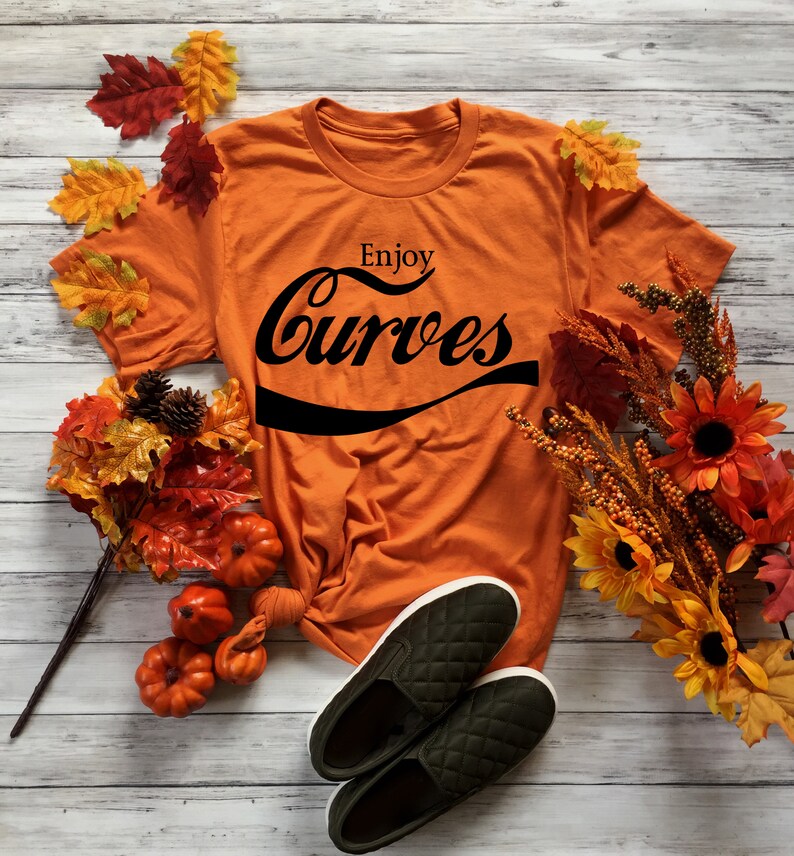 May include: Orange t-shirt with the text "Enjoy Curves" in a black retro font. The shirt is laid out on a white wooden surface with fall foliage and pumpkins.