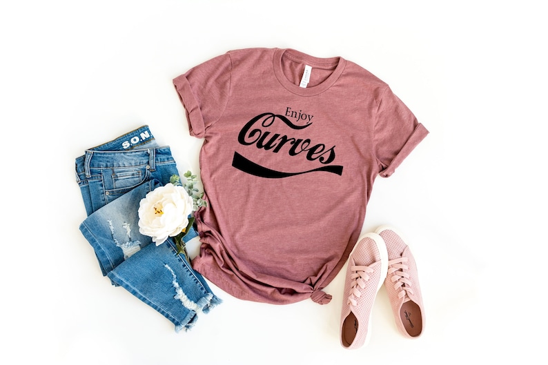 May include: A mauve t-shirt with the text "Enjoy Curves" in a black retro font. The shirt is styled with blue jeans and pink sneakers.