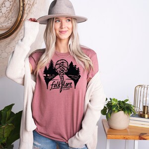 May include: A dusty rose t-shirt with a black graphic of a braided hairstyle, trees, and the word "folklore" with "Est. 2020" below it. The model is wearing a gray hat and a cream-colored cardigan.