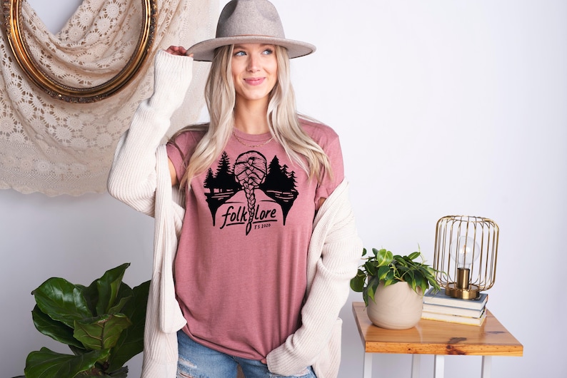 May include: A dusty rose t-shirt with a black graphic of a braided hairstyle, trees, and the word "folklore" with "Est. 2020" below it. The model is wearing a gray hat and a cream-colored cardigan.