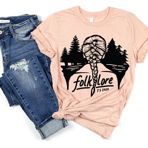 May include: A light pink t-shirt with a black graphic of a woman's braided hair, trees, and the word "folklore" with "T.S. 2020" below. The shirt is paired with blue denim jeans.