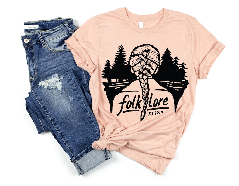 May include: A light pink t-shirt with a black graphic of a woman's braided hair, trees, and the word "folklore" with "T.S. 2020" below. The shirt is paired with blue denim jeans.