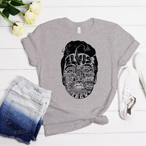May include: A gray t-shirt with a black and white illustration of Elvis Presley's face. The illustration is made up of words related to Elvis's music and career.