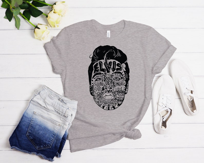 May include: A gray t-shirt with a black and white illustration of Elvis Presley's face. The illustration is made up of words related to Elvis's music and career.