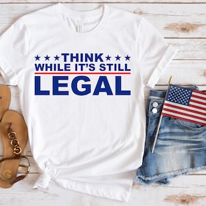 以下が含まれることがあります： 白いTシャツに「THINK WHILE IT'S STILL LEGAL」という文字が青と赤の文字で書かれています。Tシャツは木製の表面に置かれ、サンダルと小さなアメリカ国旗が付いたデニムのショートパンツが置かれています。
