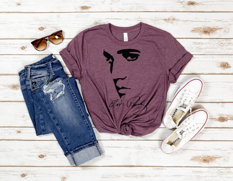 May include: A maroon t-shirt with a black silhouette of Elvis Presley's face and signature. The shirt is laid out on a white wooden surface with a pair of jeans, sunglasses, and white sneakers.