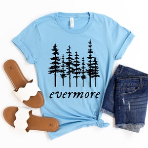 May include: A light blue t-shirt with a black tree graphic and the word "evermore". The shirt is paired with denim shorts and white sandals with brown soles. The items are arranged on a white surface.