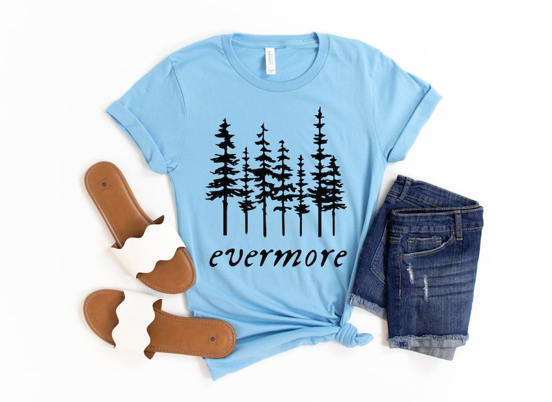 May include: A light blue t-shirt with a black tree graphic and the word "evermore". The shirt is paired with denim shorts and white sandals with brown soles. The items are arranged on a white surface.