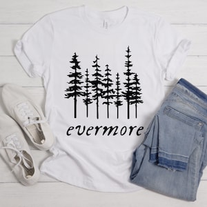 May include: White t-shirt with a black graphic of pine trees and the word "evermore" in a script font. The shirt is paired with light blue jeans and white canvas shoes, all arranged on a light wood surface.