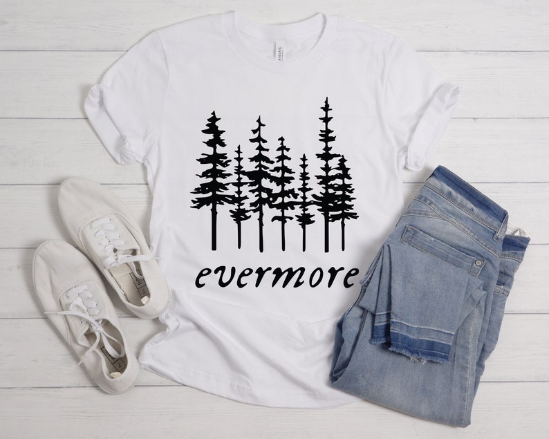 May include: White t-shirt with a black graphic of pine trees and the word "evermore" in a script font. The shirt is paired with light blue jeans and white canvas shoes, all arranged on a light wood surface.