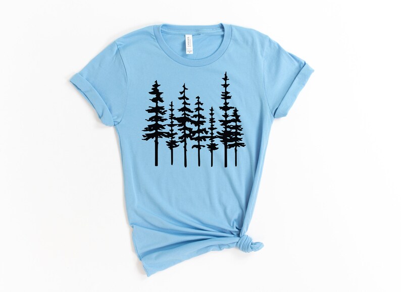 May include: Light blue t-shirt featuring a black graphic of several stylized pine trees. The shirt has short sleeves and a crew neck. The bottom of the shirt is tied in a knot.