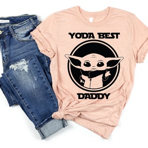 K&ouml;nnte beinhalten: Ein pfirsichfarbenes T-Shirt mit einem schwarzen Kreis, der ein Cartoon-Bild von Baby Yoda enth&auml;lt. Der Text "YODA BEST DADDY" ist in Schwarz auf das Shirt gedruckt.