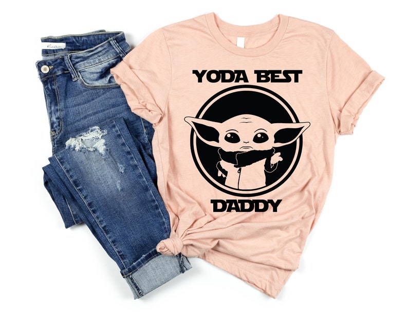 K&ouml;nnte beinhalten: Ein pfirsichfarbenes T-Shirt mit einem schwarzen Kreis, der ein Cartoon-Bild von Baby Yoda enth&auml;lt. Der Text "YODA BEST DADDY" ist in Schwarz auf das Shirt gedruckt.