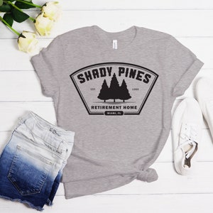 Może przedstawiać: Szara koszulka z czarnym nadrukiem logo, na kt&oacute;rym widnieje napis "Shady Pines Retirement Home" z motywem sosny i tekstem "Est. 1985".