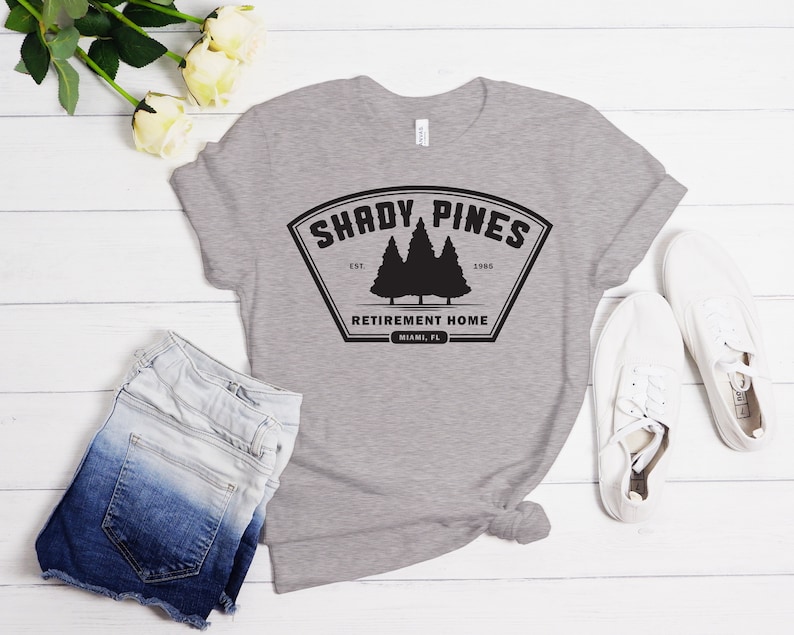 Może przedstawiać: Szara koszulka z czarnym nadrukiem logo, na kt&oacute;rym widnieje napis "Shady Pines Retirement Home" z motywem sosny i tekstem "Est. 1985".