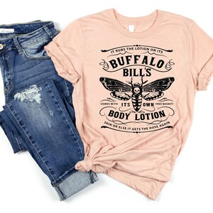 May include: A peach-colored t-shirt with a black graphic of a moth with a skull and crossbones. The graphic says "It rubs the lotion on its Buffalo Bill's. Comes with its own free basket! Body Lotion. Skin or else it gets the hose again."