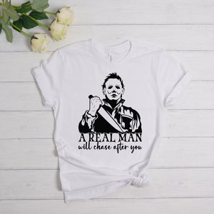 K&ouml;nnte beinhalten: Wei&szlig;es T-Shirt mit einer schwarz-wei&szlig;en Grafik eines Mannes, der ein Messer h&auml;lt. Der Text "A REAL MAN will chase after you" ist unter der Grafik gedruckt.