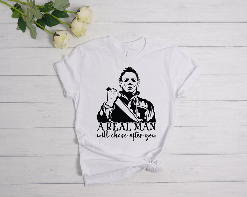 K&ouml;nnte beinhalten: Wei&szlig;es T-Shirt mit einer schwarz-wei&szlig;en Grafik eines Mannes, der ein Messer h&auml;lt. Der Text "A REAL MAN will chase after you" ist unter der Grafik gedruckt.