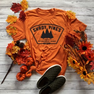 Może przedstawiać: Pomarańczowy T-shirt z czarną grafiką logo, kt&oacute;re brzmi "Shady Pines Retirement Home, Miami, FL" z czarną sylwetką trzech sosen. Logo znajduje się w czarnym kształcie tarczy z białym obramowaniem. T-shirt leży na białym drewnianym tle z jesiennymi liśćmi i dyniami.