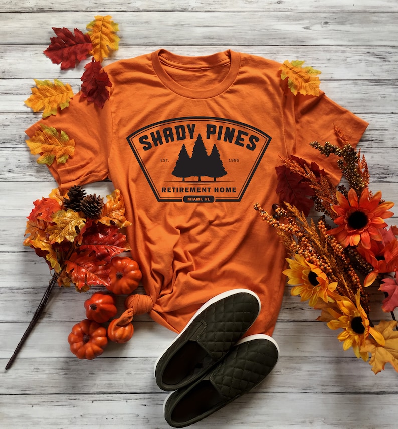 Może przedstawiać: Pomarańczowy T-shirt z czarną grafiką logo, kt&oacute;re brzmi "Shady Pines Retirement Home, Miami, FL" z czarną sylwetką trzech sosen. Logo znajduje się w czarnym kształcie tarczy z białym obramowaniem. T-shirt leży na białym drewnianym tle z jesiennymi liśćmi i dyniami.