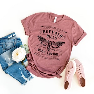 May include: A pink t-shirt with a black graphic design featuring a moth and the text "IT RUBS THE LOTION ON ITS BUFFALO BILL'S ITS OWN BODY LOTION SKIN OR ELSE IT GETS THE HOSE AGAIN". The shirt is paired with blue jeans and pink sneakers.