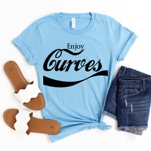 May include: A light blue t-shirt with the text "Enjoy Curves" in a black, stylized font. The shirt is paired with denim shorts and white sandals.