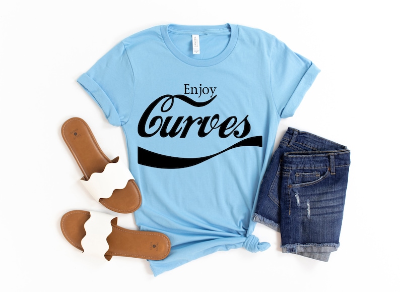 May include: A light blue t-shirt with the text "Enjoy Curves" in a black, stylized font. The shirt is paired with denim shorts and white sandals.
