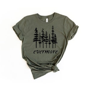 May include: Olive green t-shirt featuring a black graphic of several pine trees and the word "evermore" in a script font. The shirt has rolled-up sleeves and a slightly wrinkled texture.