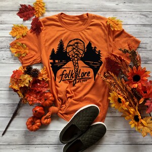 May include: Orange t-shirt with a black graphic of a braided hairstyle and a forest silhouette, with the word "folklore" and "FS 2020". The shirt is paired with green quilted slip-on shoes and autumn decorations.