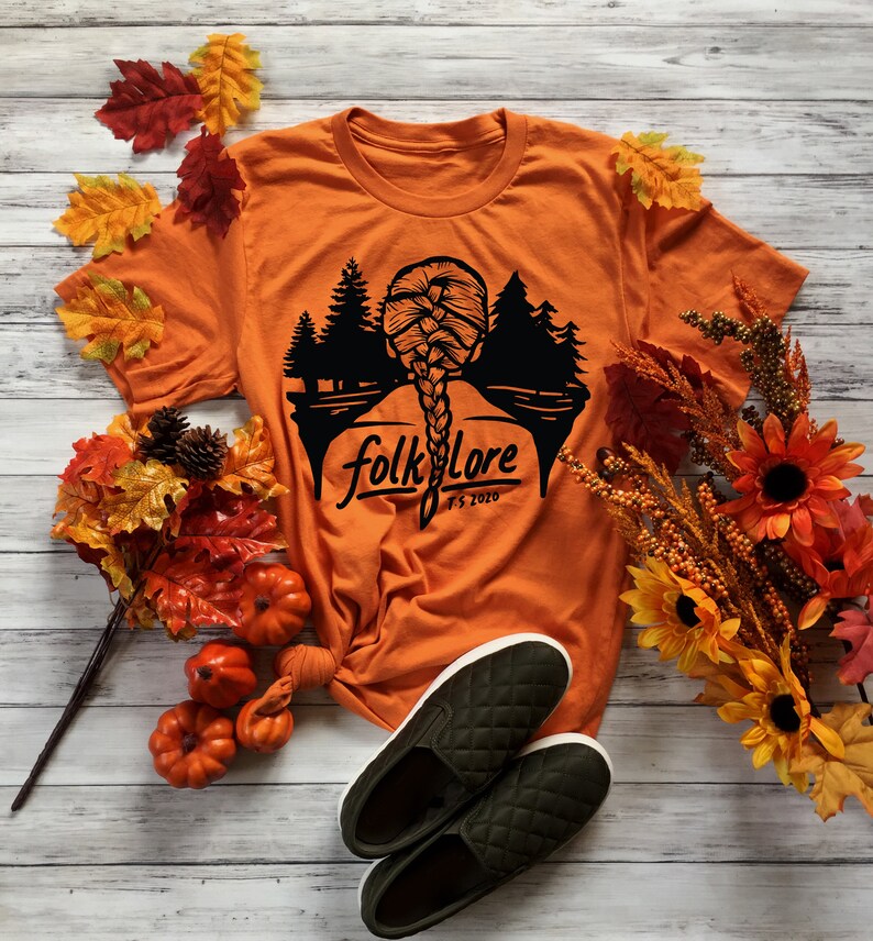 May include: Orange t-shirt with a black graphic of a braided hairstyle and a forest silhouette, with the word "folklore" and "FS 2020". The shirt is paired with green quilted slip-on shoes and autumn decorations.