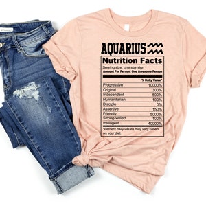 May include: A pink t-shirt with the text "Aquarius Nutrition Facts" and a list of personality traits with percentages. The text includes the symbol for the Aquarius zodiac sign.