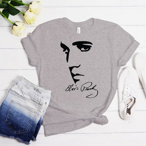 May include: A gray t-shirt with a black silhouette of Elvis Presley's face and his signature. The shirt is laid flat on a white wooden surface with a pair of white sneakers and a pair of denim shorts.