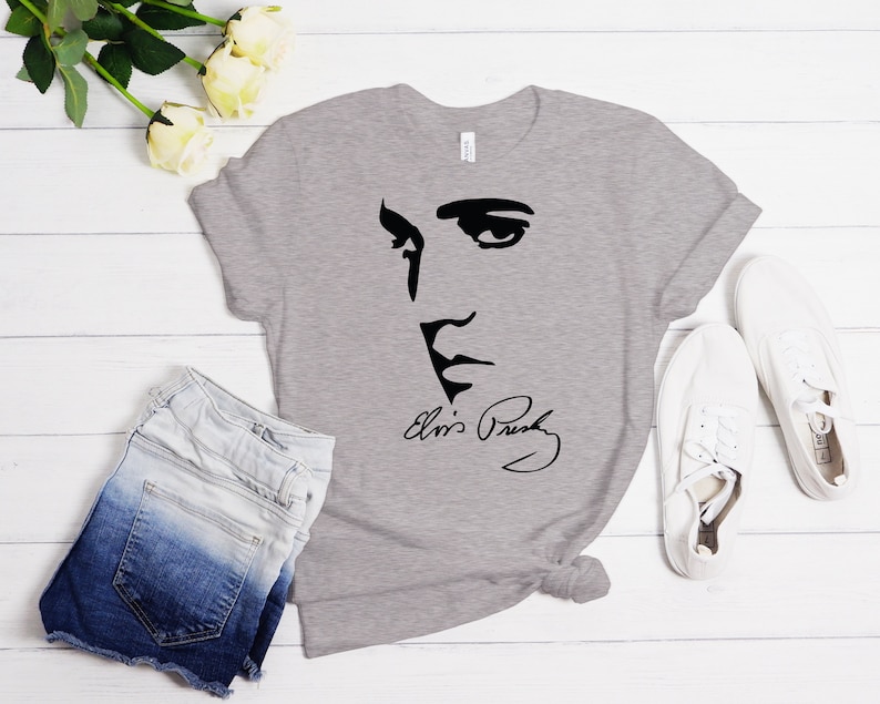 May include: A gray t-shirt with a black silhouette of Elvis Presley's face and his signature. The shirt is laid flat on a white wooden surface with a pair of white sneakers and a pair of denim shorts.
