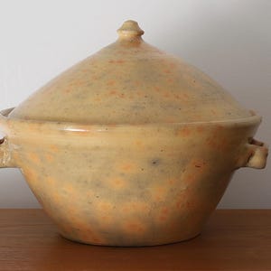 May include: A handmade, lidded ceramic casserole dish. The dish is a light yellow color with orange speckles and two side handles. The lid has a rounded shape with a small knob on top. The dish is sitting on a wooden surface.