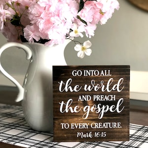 May include: A wooden sign with a dark brown stain and white lettering. The sign reads "GO INTO ALL the world AND PREACH the gospel TO EVERY CREATURE. Mark 16:15".