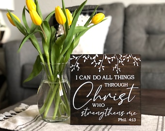 May include: A dark brown wooden sign with white lettering that reads "I can do all things through Christ who strengthens me Phil. 4:13". The sign has a white floral design at the top and is sitting on a table with a white and gray striped tablecloth.
