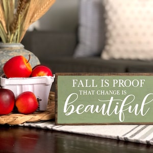 May include: A wooden sign with the text "FALL IS PROOF THAT CHANGE IS BEAUTIFUL" in white lettering. The sign is on a table with red apples, a woven basket, and a vase with wheat stalks. The background is blurred.